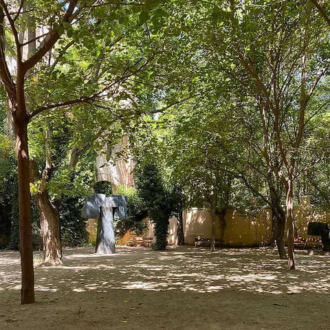 Jardín arbolado con suelo de tierra, bancos y una escultura al fondo.
