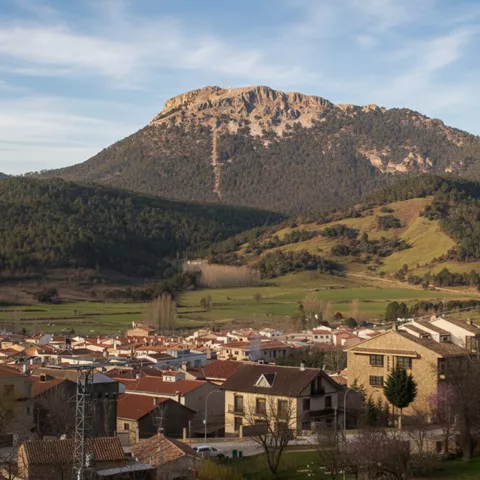Pueblo con tejados rojos al pie de una gran montaña boscosa bajo cielo claro.