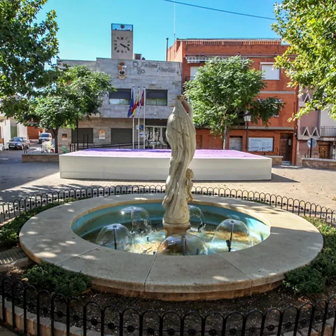 Fuente circular con escultura central en una plaza arbolada.