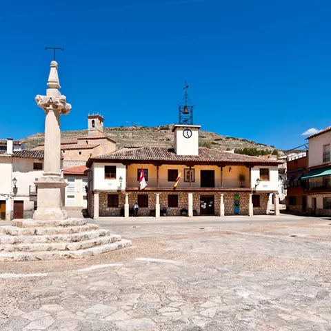 Plaza empedrada con edificio porticado y columna monumental bajo cielo azul.