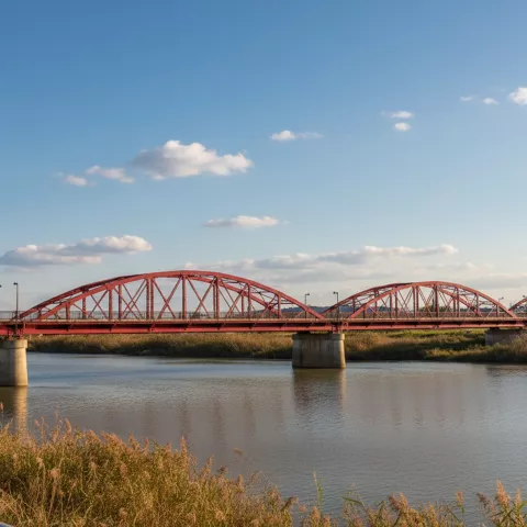 Puente metálico rojo de arcos sobre un río, con cielo azul y nubes.
