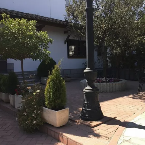 Patio exterior con farola, jardineras y mesas entre árboles.