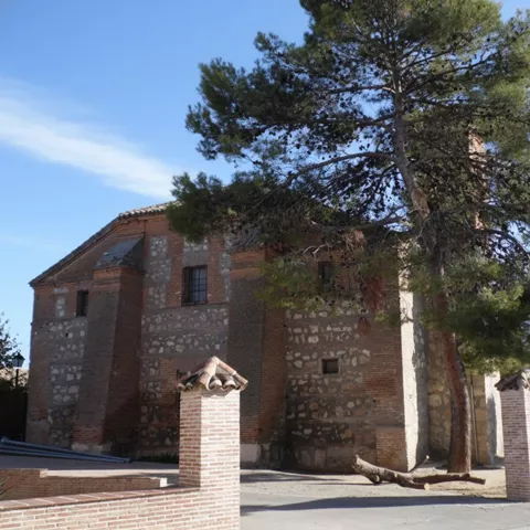 Vista de un gran edificio de piedra y ladrillo parcialmente oculto por un gran pino maduro, con una entrada flanqueada por pilares de ladrillo bajos, bajo un cielo azul con nubes ligeras.