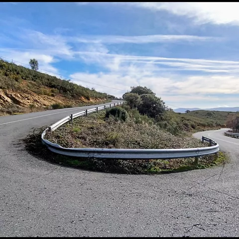 Curva cerrada de carretera de montaña con quitamiedos y cielo despejado.