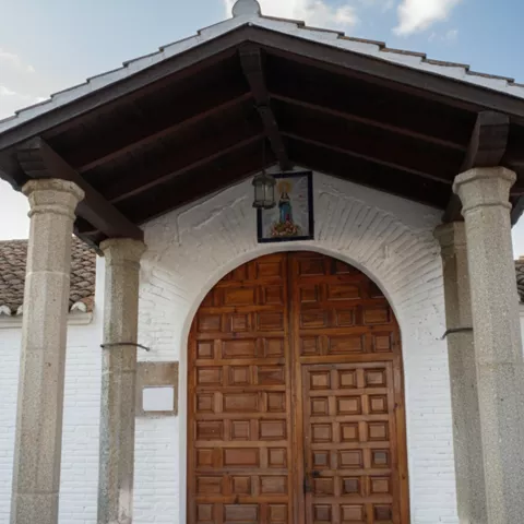 Una puerta de madera robusta bajo un arco blanco, protegida por un porche con columnas de piedra y techo de vigas de madera.