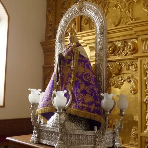 Imagen religiosa en andas con manto morado, rodeada de candelabros ante retablo dorado.