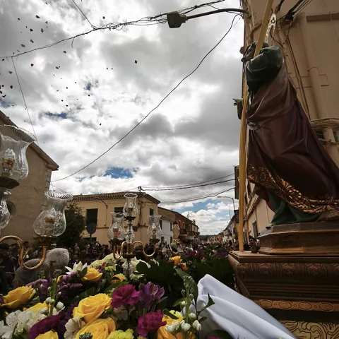 Paso procesional con imagen religiosa entre flores y faroles en una calle llena de público.