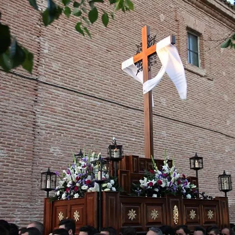 Cruz de madera adornada con tela blanca y flores en un paso procesional.