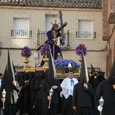 Procesión con nazarenos de negro y paso de Jesús con la cruz adornado con flores moradas.