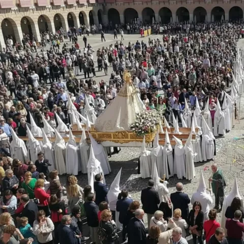 Procesión multitudinaria en plaza porticada con nazarenos vestidos de blanco y pasos florales.