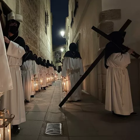 Penitentes con túnicas blancas y capirotes negros portando faroles y una cruz en una calle estrecha de noche.