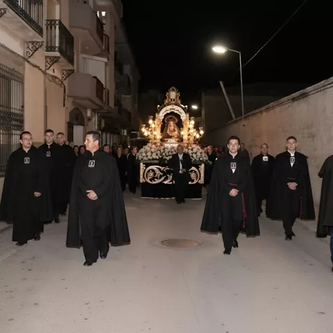 Procesión nocturna con imagen religiosa iluminada y participantes vestidos de negro.