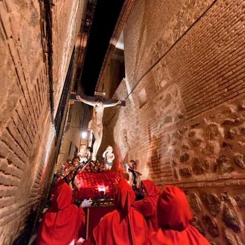 Procesión nocturna por una calle estrecha, con imagen religiosa iluminada y participantes con túnicas.