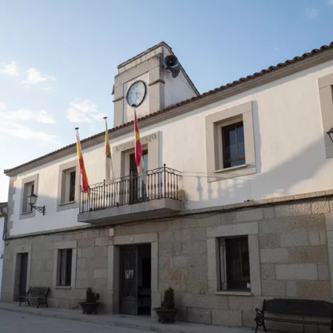 Plano medio de la entrada principal de un edificio público blanco, con tres banderas colgando de un balcón y un gran reloj central en la parte superior.