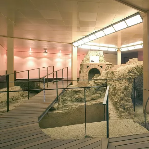 Sala musealizada con pasarelas y barandillas sobre restos de piedra y muros antiguos.