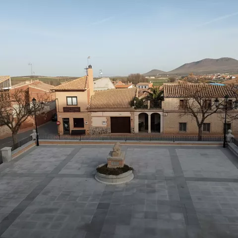 Plaza de un pueblo con una escultura de piedra en el centro, rodeada de casas de arquitectura tradicional y colinas bajas al fondo.