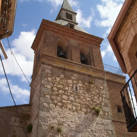 Vista en ángulo bajo de una torre de piedra y ladrillo con dos campanas visibles en sus arcos superiores bajo un cielo azul con nubes.