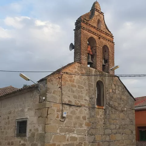 Primer plano de una espadaña de ladrillo con dos campanas sobre una estructura de piedra rústica, con farolas modernas y cables eléctricos cruzando la escena.