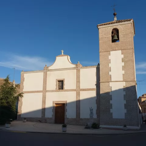 Fachada de una iglesia sencilla de color blanco con detalles en ladrillo visto y una torre campanario lateral coronada por una cruz metálica.