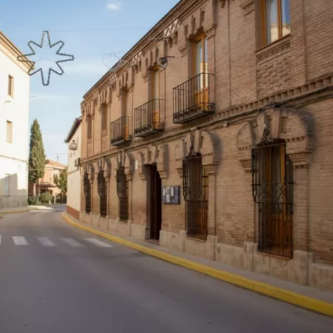 Una calle de pueblo con bordillos amarillos que se curva hacia la distancia, flanqueada a la derecha por un gran edificio de ladrillo con un patrón detallado sobre las ventanas de la planta baja.