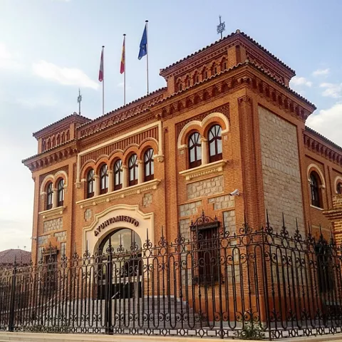 Primer plano de una fachada de ladrillo rojo y piedra, con un gran arco de entrada, balcones de hierro forjado y tres banderas.