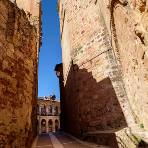 Calle estrecha entre muros de piedra con torres visibles al fondo.