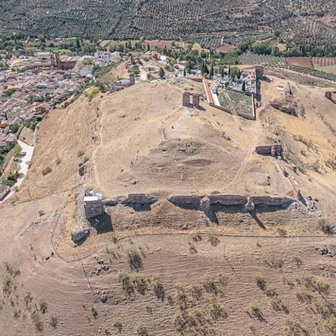 Vista aérea de un cerro con restos de murallas y ruinas de piedra junto a un núcleo urbano y campos de cultivo.
