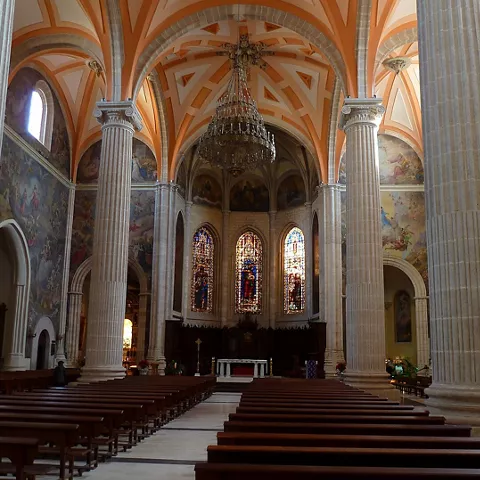 Nave central de iglesia con columnas, vitrales y bancos alineados.