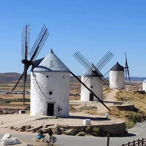 Molinos de viento alineados junto a una fortaleza sobre un paisaje rural.