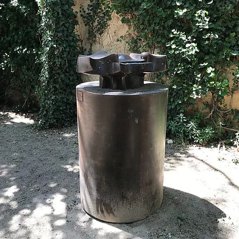 Escultura geométrica de metal sobre base cilíndrica en un jardín sombreado.