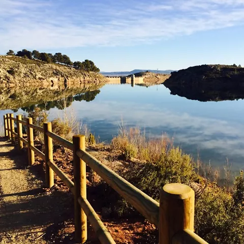 Sendero de madera junto a un embalse con reflejos del paisaje en el agua.