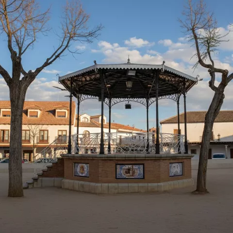 Quiosco metálico rodeado de árboles en plaza pavimentada con edificios tradicionales.