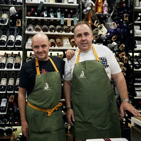 Dos hombres con delantal posan frente a estanterías llenas de botellas de vino.