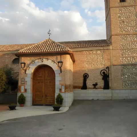 La entrada principal de una iglesia de ladrillo con un arco de piedra alrededor de una puerta de madera, un campanario alto y decoraciones metálicas de una escena de natividad en la pared lateral.