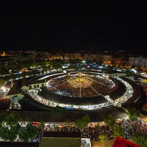 Vista aérea nocturna desde gran altura de un recinto circular iluminado, rodeado de zonas verdes y edificios.