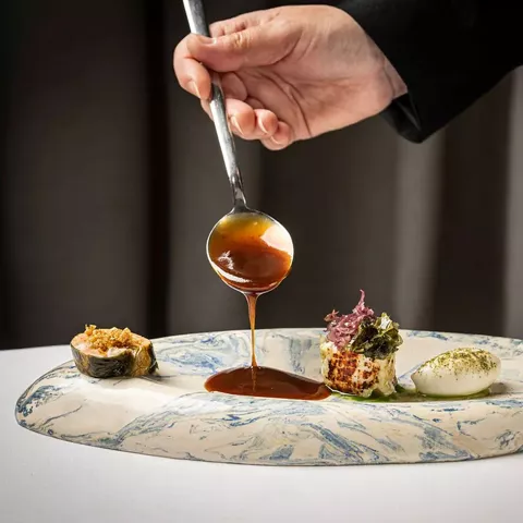 Mano vertiendo salsa sobre plato de alta cocina con presentaciones minimalistas.