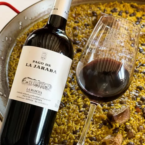 Botella y copa de vino tinto delante de una paella en una sartén grande.