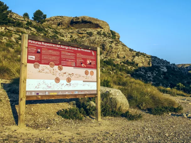 Panel informativo en un paraje rocoso y seco, con sendero y matorral.