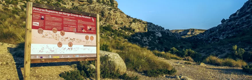 Panel informativo en un paraje rocoso y seco, con sendero y matorral.