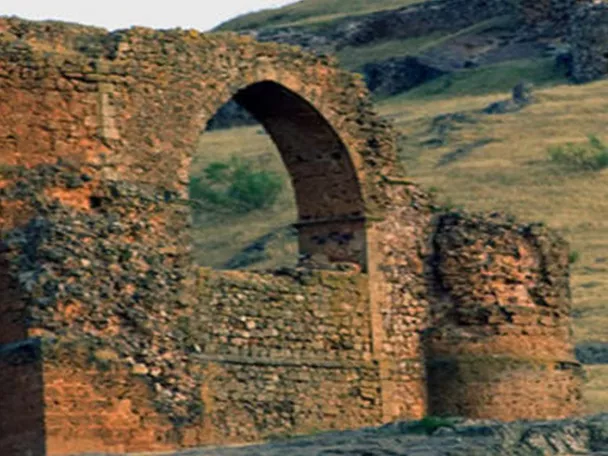 Restos de construcción de piedra con arco central sobre una ladera cubierta de pasto.