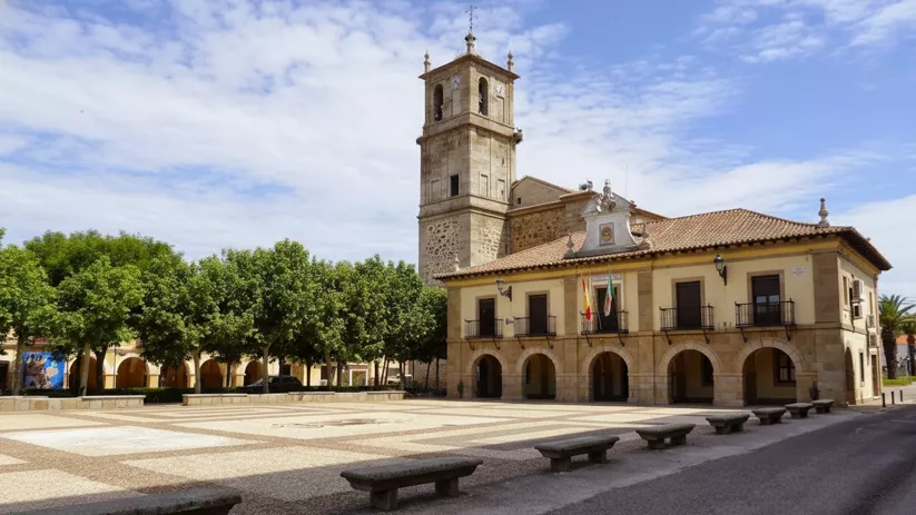 Plaza porticada con edificio institucional y torre de piedra al fondo.