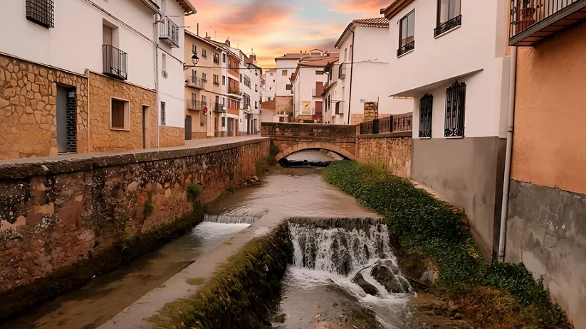 Calle estrecha con casas blancas junto a un canal de agua y un pequeño puente al atardecer.