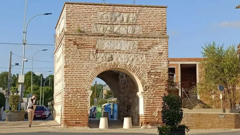 Arco histórico de ladrillo en una rotonda con peatones y contenedores.