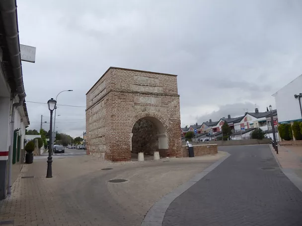 Arco de ladrillo y piedra en una plaza urbana con cielo nublado.