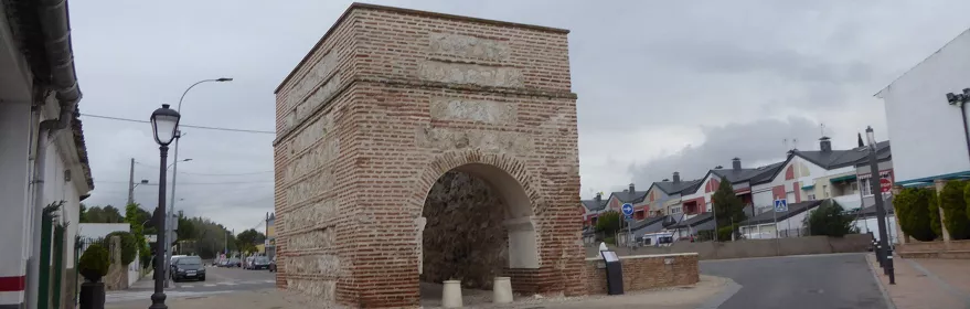 Arco de ladrillo y piedra en una plaza urbana con cielo nublado.