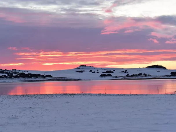 Atardecer invernal con cielo rosado reflejado en un lago rodeado de nieve.