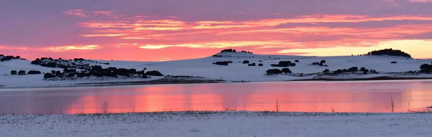Atardecer invernal con cielo rosado reflejado en un lago rodeado de nieve.