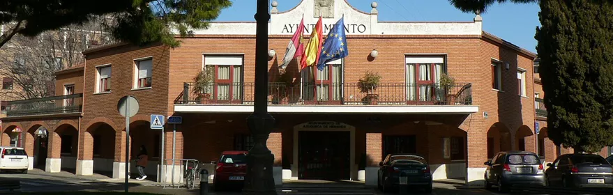 Edificio institucional con reloj y banderas en una plaza arbolada