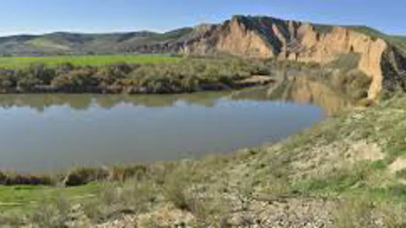 Lago tranquilo con laderas verdes y cortados de tierra rojiza al fondo.