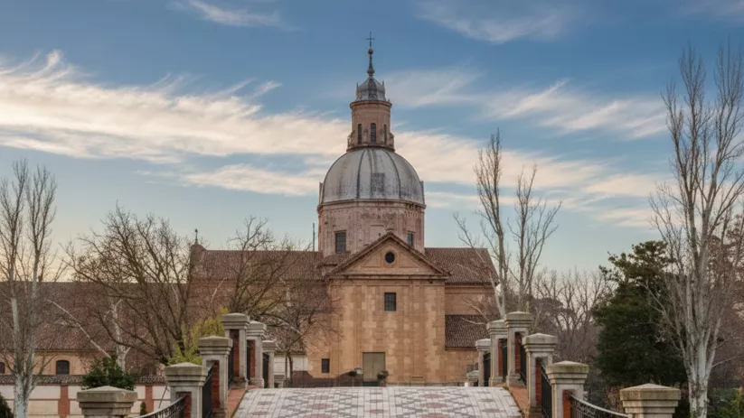 Puente empedrado alineado hacia un templo de piedra con gran cúpula y cielo despejado.